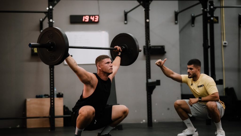 A muscular man performing a weightlifting squat with a coach guiding him indoors.