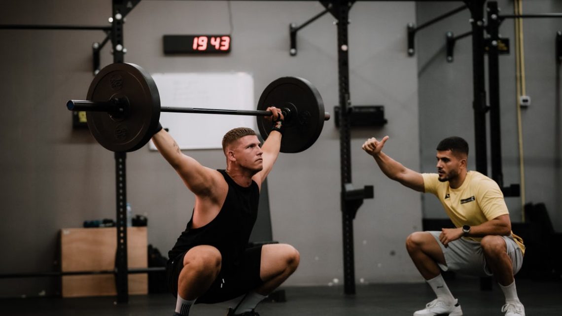 A muscular man performing a weightlifting squat with a coach guiding him indoors.