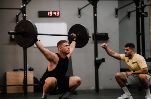 A muscular man performing a weightlifting squat with a coach guiding him indoors.