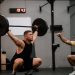 A muscular man performing a weightlifting squat with a coach guiding him indoors.