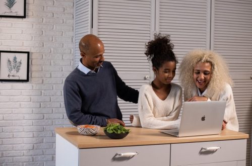 Positive African American parent and daughter surfing modern netbook while standing at wooden kitchen counter with fresh ingredients at home