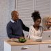Positive African American parent and daughter surfing modern netbook while standing at wooden kitchen counter with fresh ingredients at home