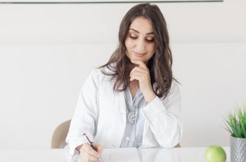 A female nutritionist in a lab coat writing notes with an apple and plant on the desk.